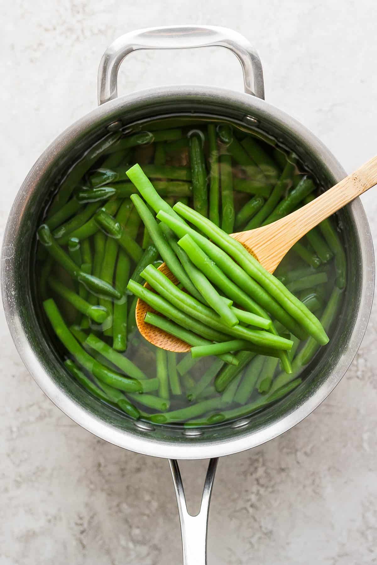 Boiled green beans in a sauce pan filled with water with a wooden slotted spoon pulling a few out.