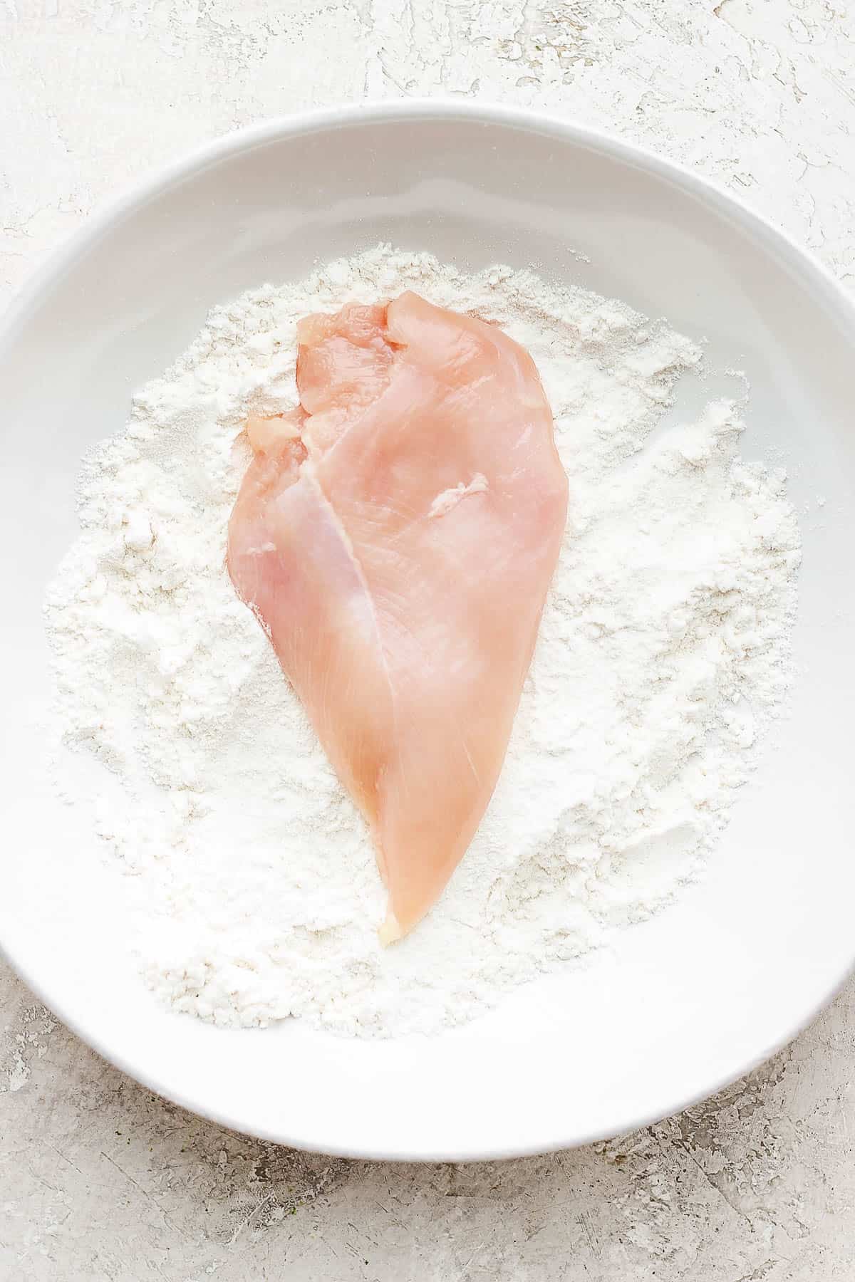 Raw chicken cutlet in a shallow bowl of flour.