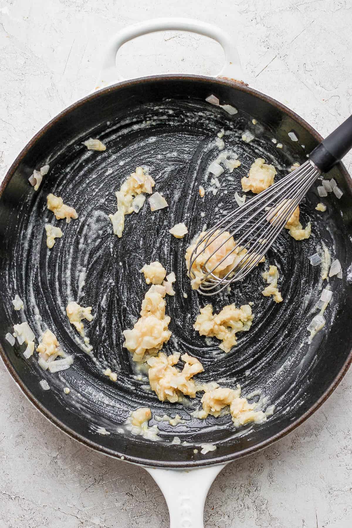 A whisk mixing in flour to the onion, butter, and garlic mixture.
