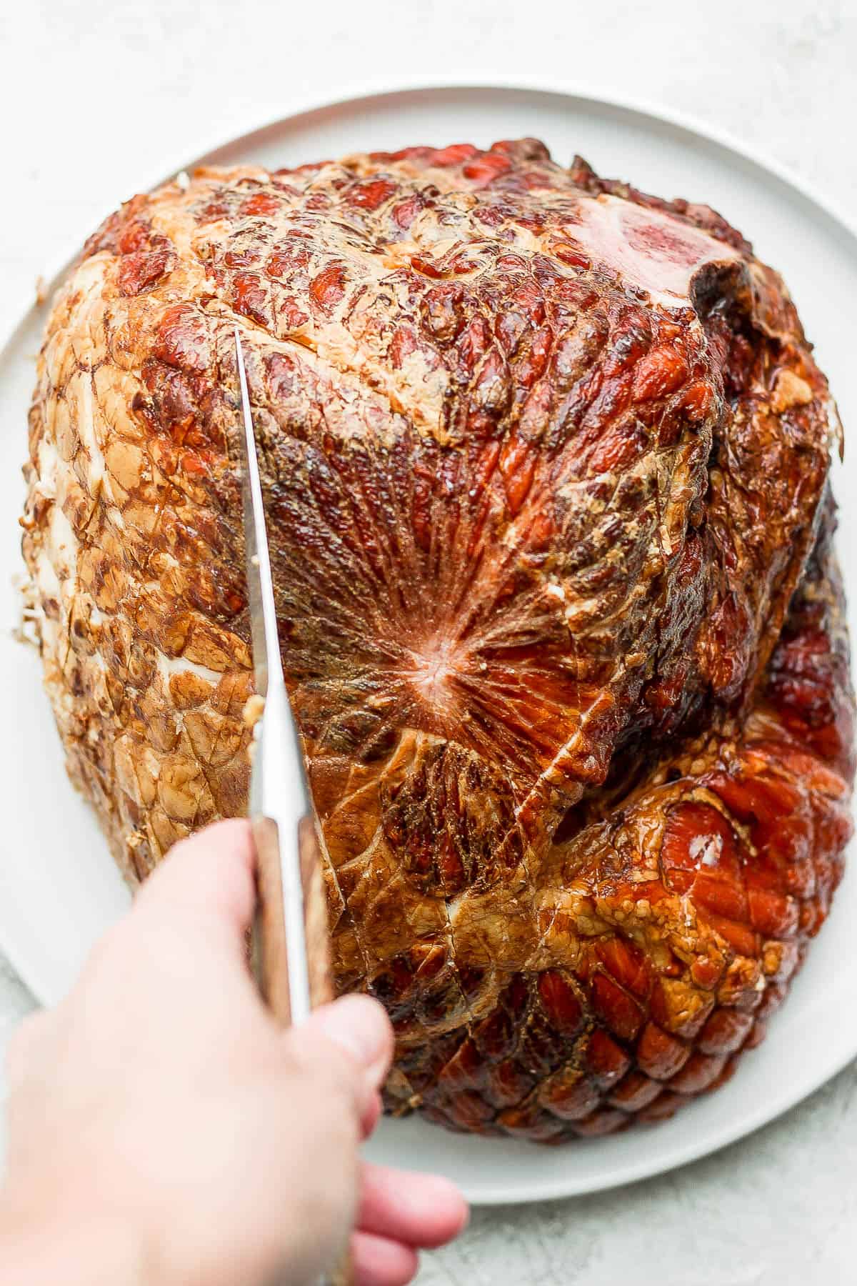 A ham on a white plate being scored with a pairing knife.