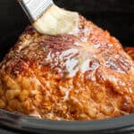 Close up of a ham in a crockpot and someone brushing brown sugar glaze on top.