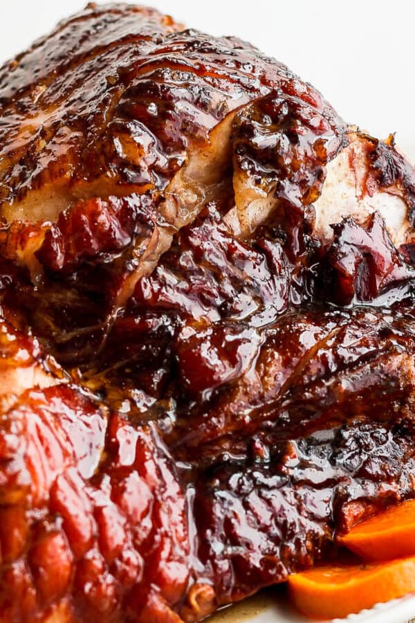 Side view of a crockpot ham on a plate with glaze on top and oranges and herbs on the plate around it.