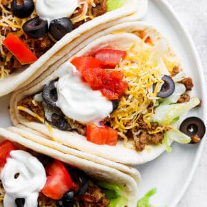 Top shot of three soft shell ground beef tacos with sour cream, olives, tomatoes and cheese on top.