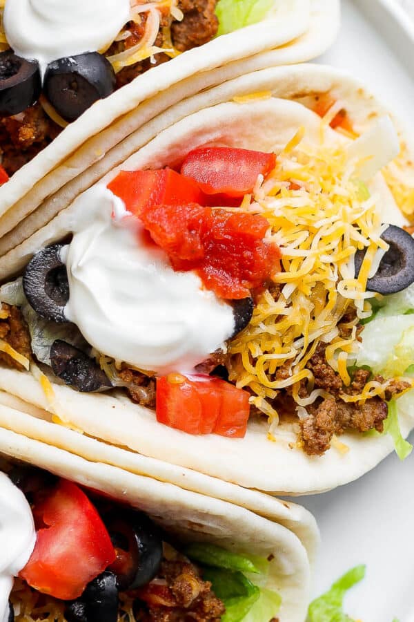 Top shot of three soft shell ground beef tacos with sour cream, olives, tomatoes and cheese on top.