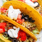 Three hard shell ground beef tacos next to each other with sour cream, olives, tomatoes and cheese on top.
