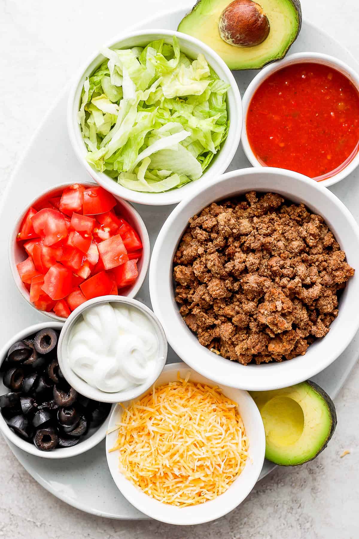 A serving platter with seven small containers each holding individual ingredients; shredded romaine lettuce, taco sauce, ground beef, chopped tomatoes, sour cream, sliced black olives, shredded cheese, and avocado. 