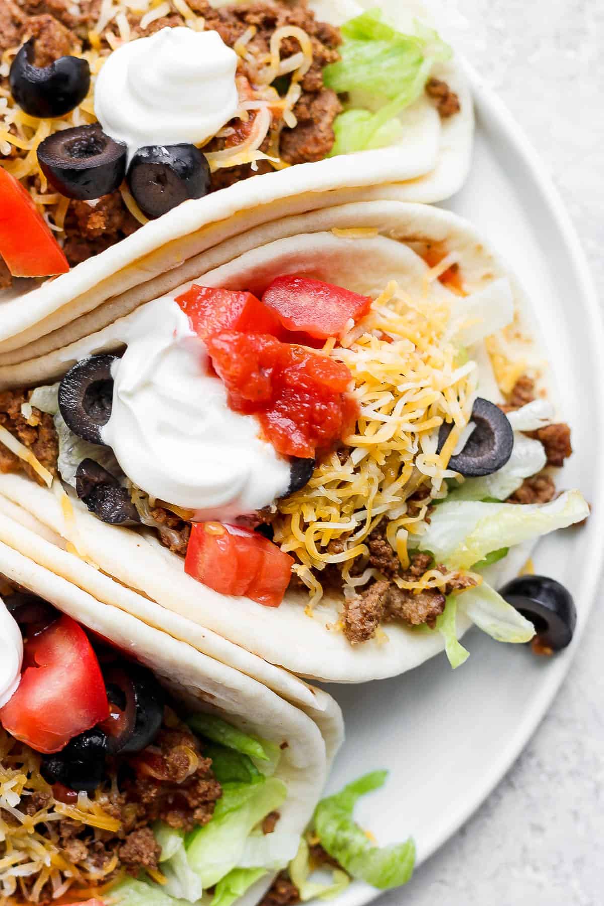 A close up of a ground beef taco topped with shredded cheese, sliced olives, taco sauce, and sour cream.