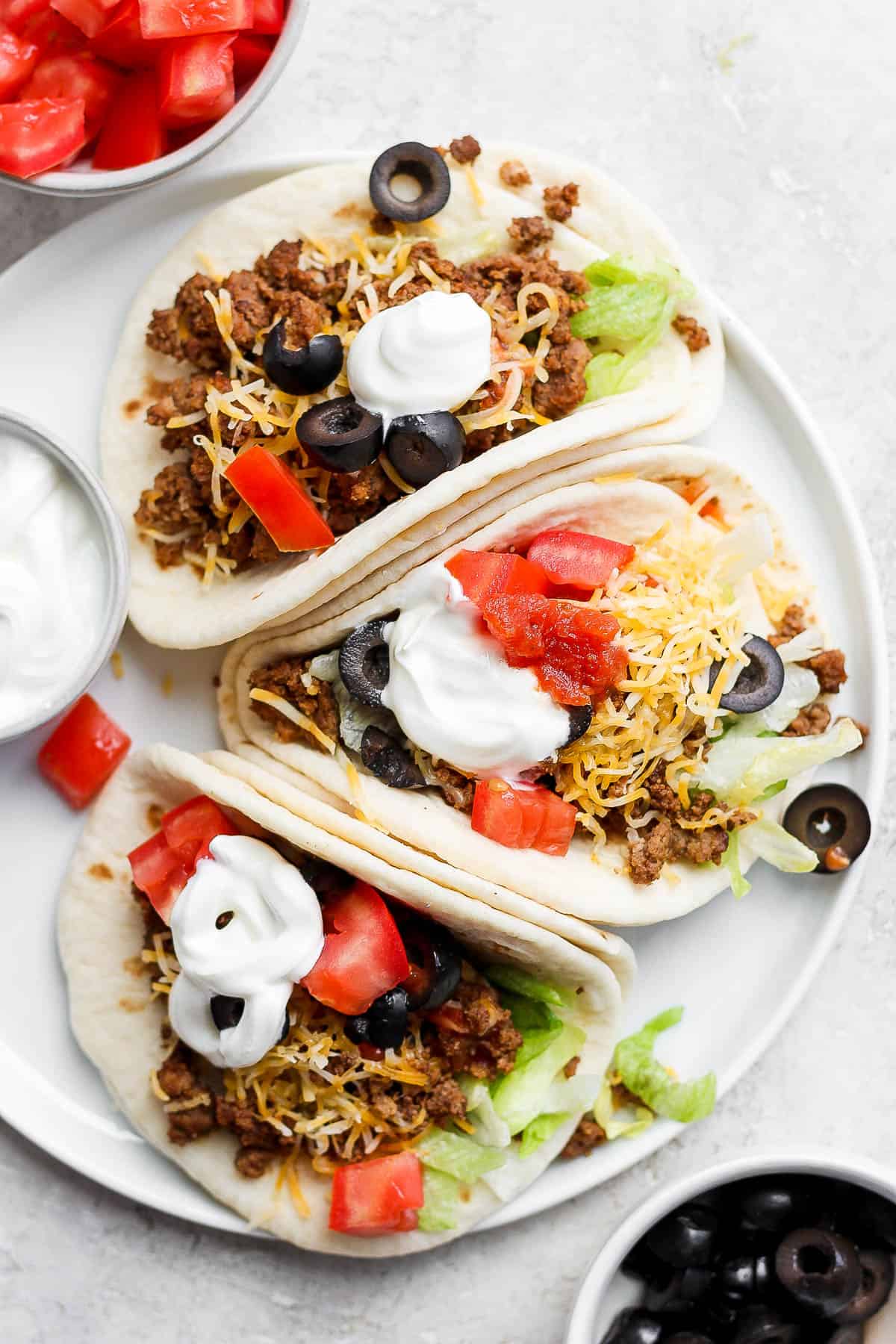 Three ground beef tacos on a plate topped with shredded cheese, chopped tomatoes, sour cream, and sliced black olives. 