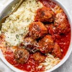 Bowl of Italian Meatballs in red sauce with parmesan orzo and fresh oregano leaves on top.
