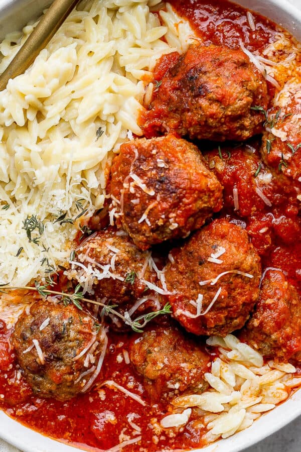 Bowl of Italian Meatballs in red sauce with parmesan orzo and fresh oregano leaves on top.