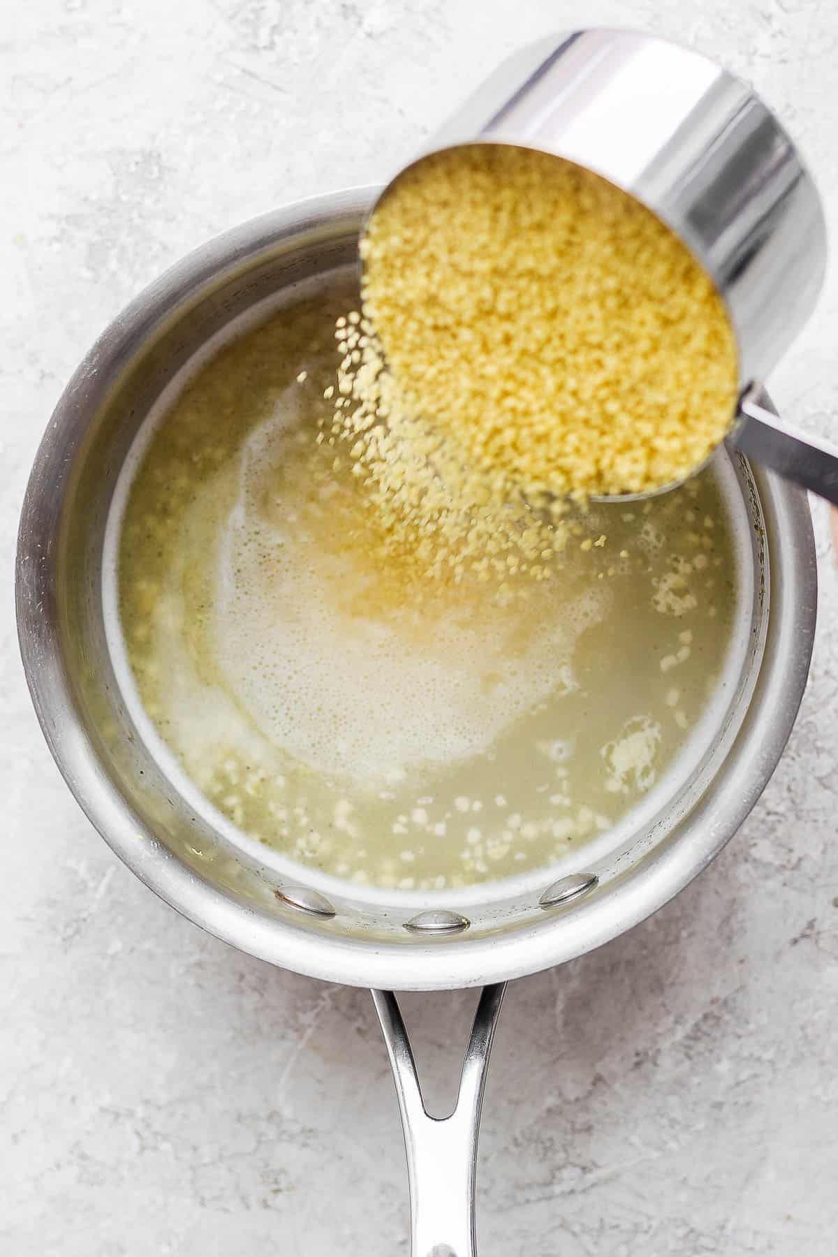 A cup of raw couscous being poured into the water mixture in the sauce pan.