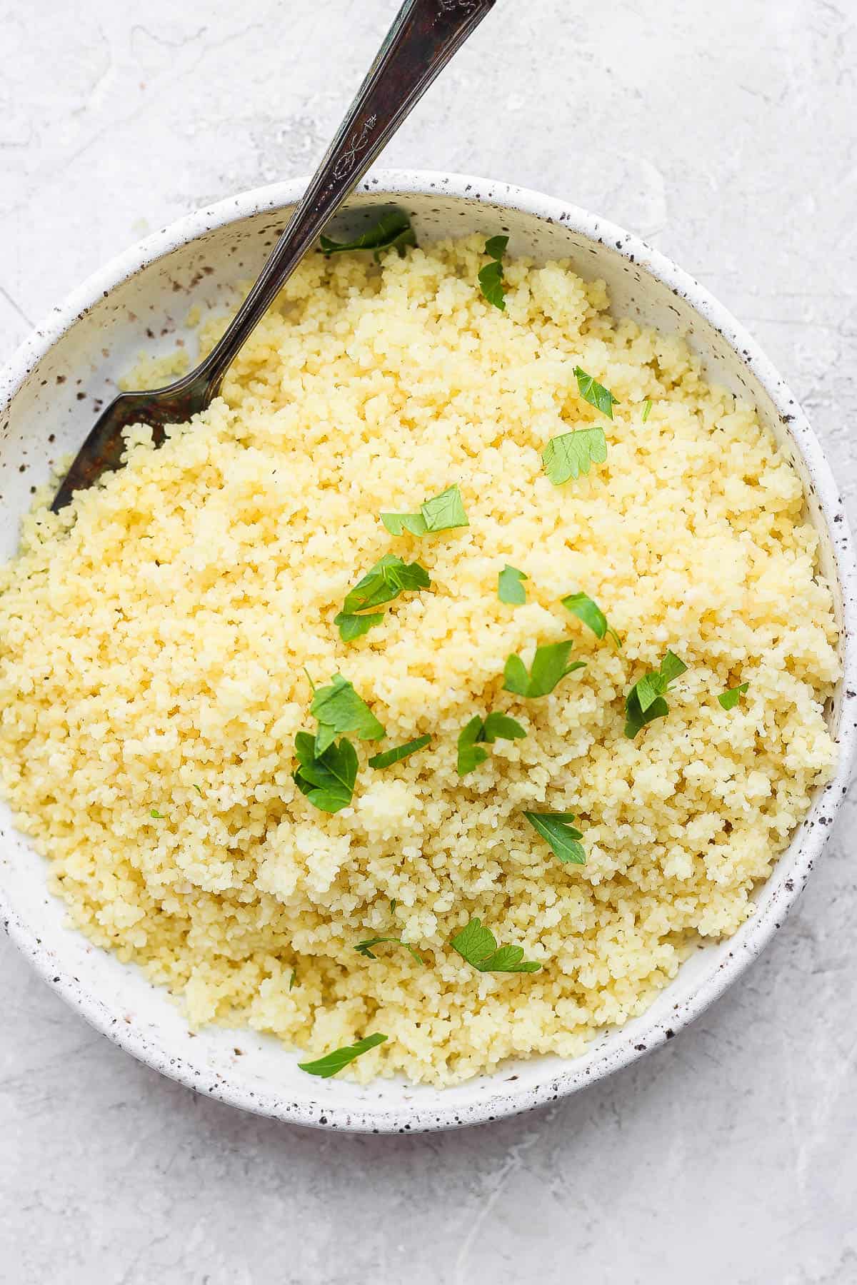 Cooked couscous in a serving bowl and sprinkled with chopped parsley.  A fork is off to the side of the bowl.