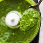 A spoon with mint chimichurri on it held over a small food processor also filled with mint chimichurri.