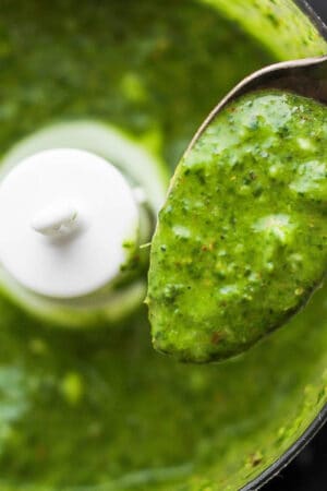 A spoon with mint chimichurri on it held over a small food processor also filled with mint chimichurri.