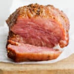 A smoked corned beef sitting on a piece of parchment on top of a wooden board with one slice sitting in from of the rest of the roast.