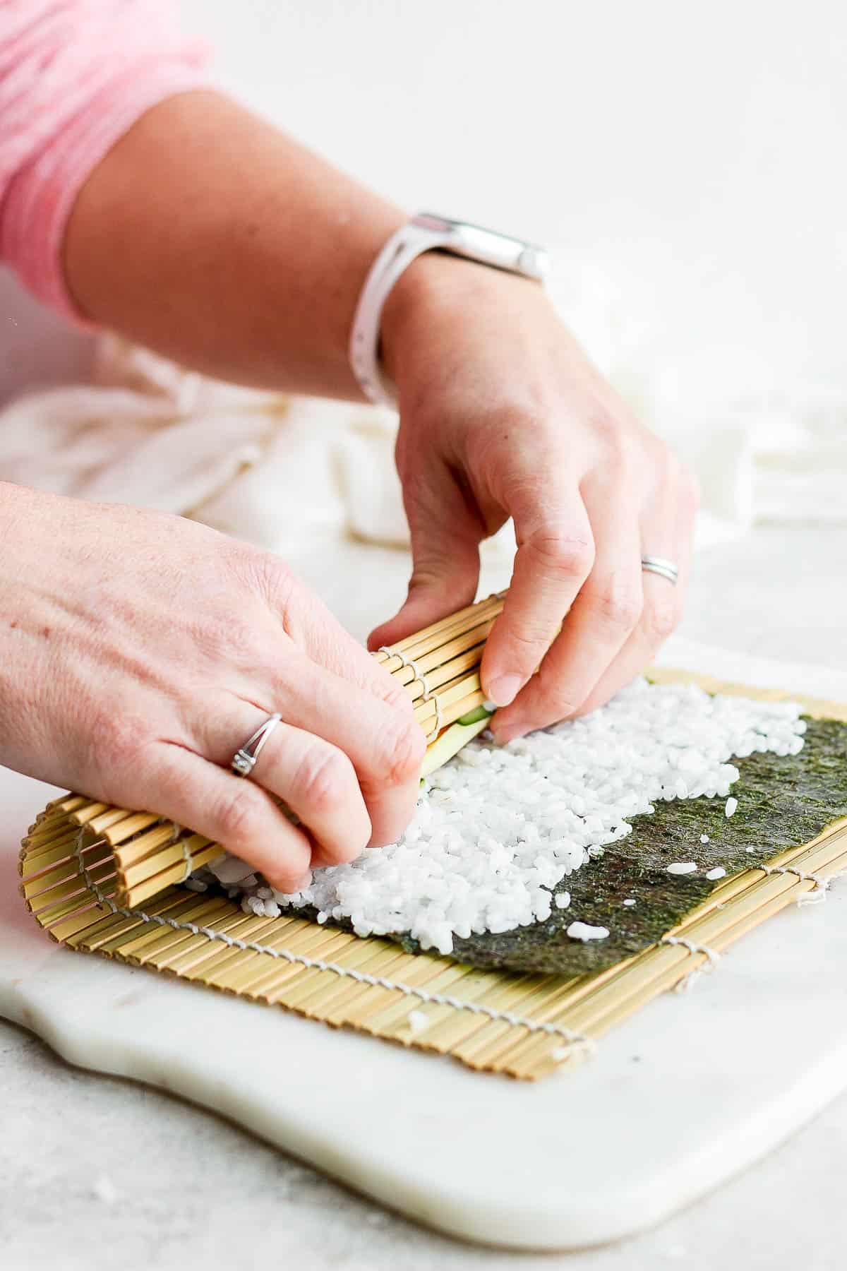 Two hands pulling the bamboo sheet and filled nori sheet over the filling ingredients.