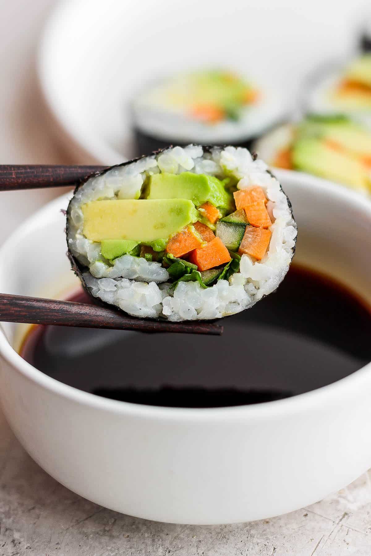 Chopsticks holding a piece of avocado roll over a small bowl of soy sauce.
