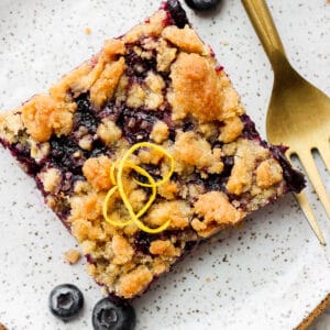 A blueberry crumb bar on a plate with a piece of lemon zest on top next to a fork and a few blueberries.