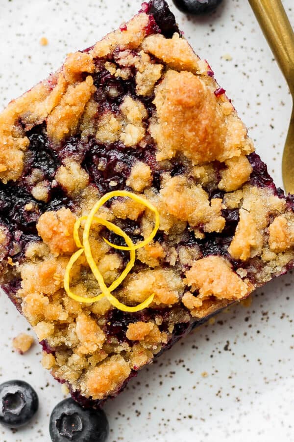 A blueberry crumb bar on a plate with a piece of lemon zest on top next to a fork and a few blueberries.