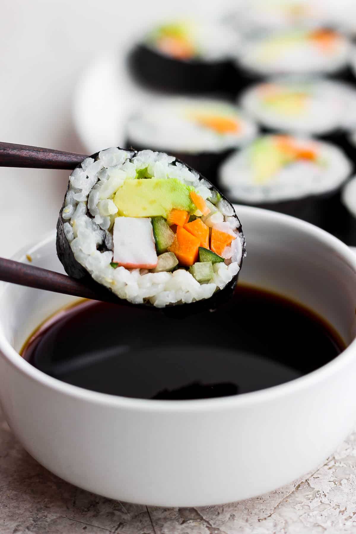 Chopsticks holding a piece of a California roll over a bowl of soy sauce.