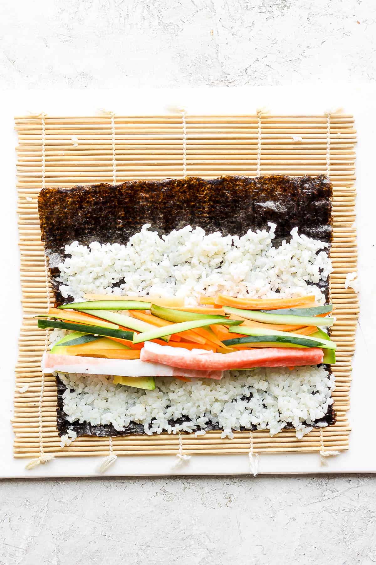 A nori sheet on a bamboo sheet with rice spread on top and fillings added.