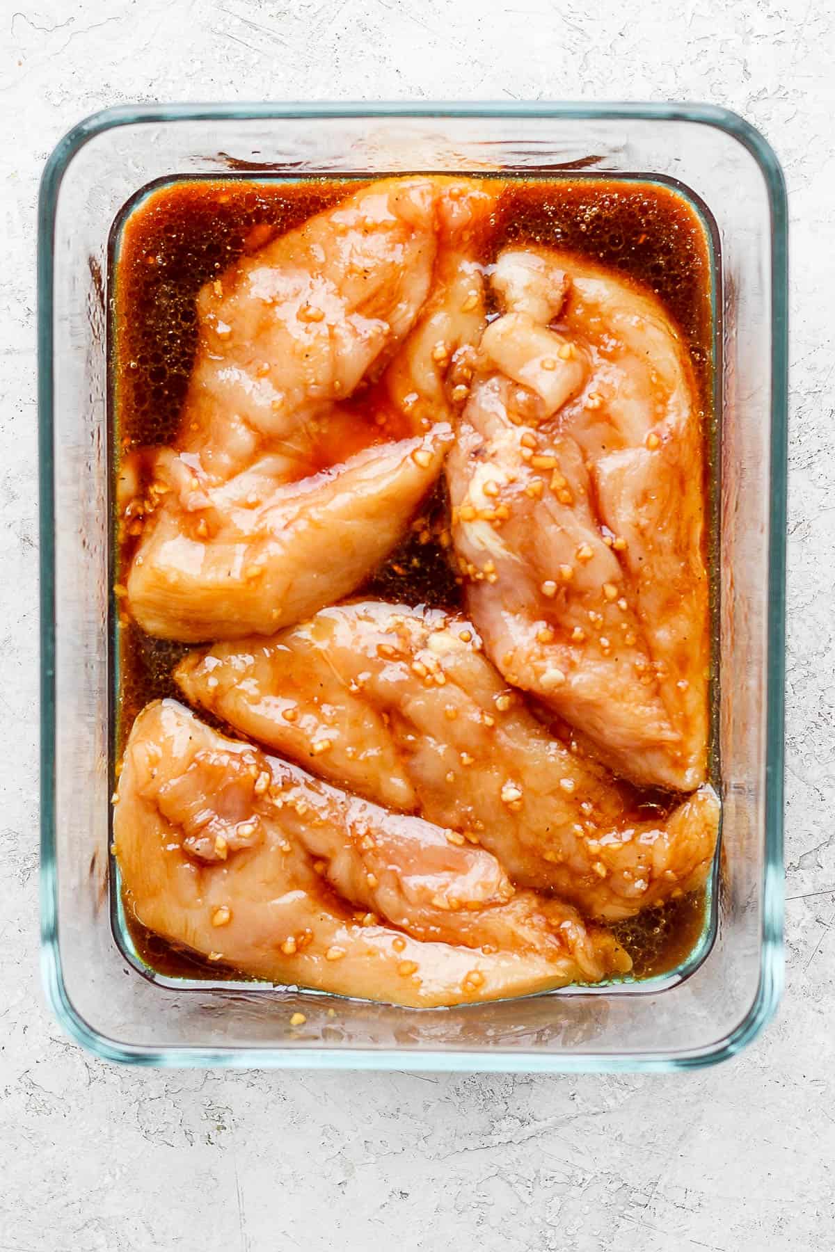 four chicken breasts topped with marinade in a glass dish.