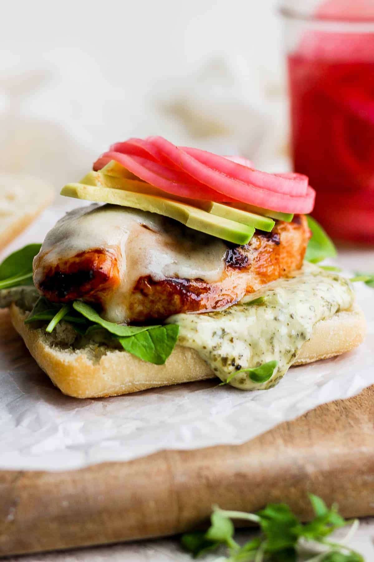 A focaccia bun topped with pesto aioli, arugula, grilled chicken breast, cheese, avocado slices, and pickled onions.