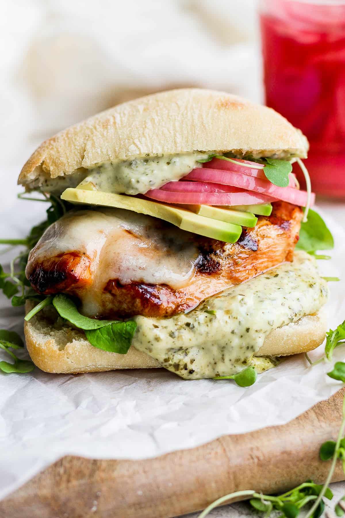 A focaccia bun topped with pesto aioli, arugula, grilled chicken breast, cheese, avocado slices, and pickled onions and topped with the other piece of focaccia bun.