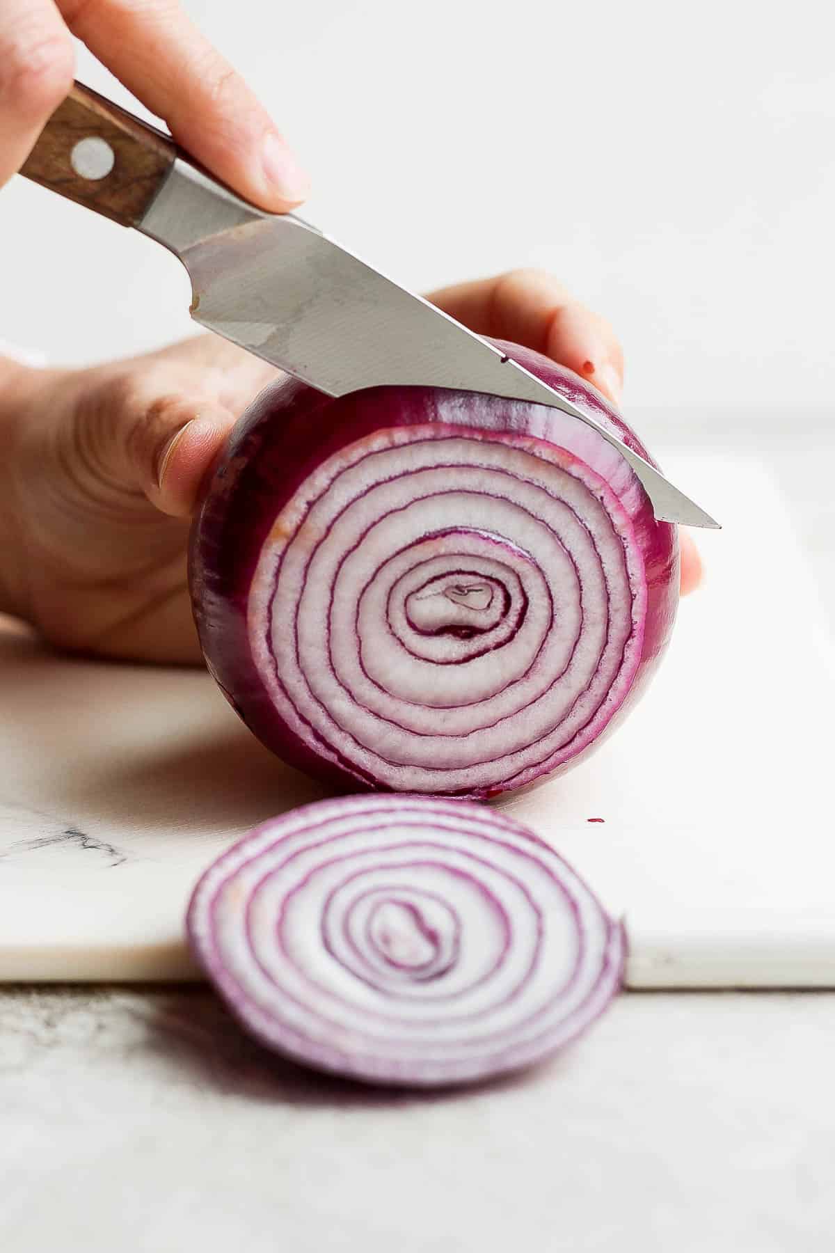 A sharp knife cutting a large red onion into thick slices on a cutting board.