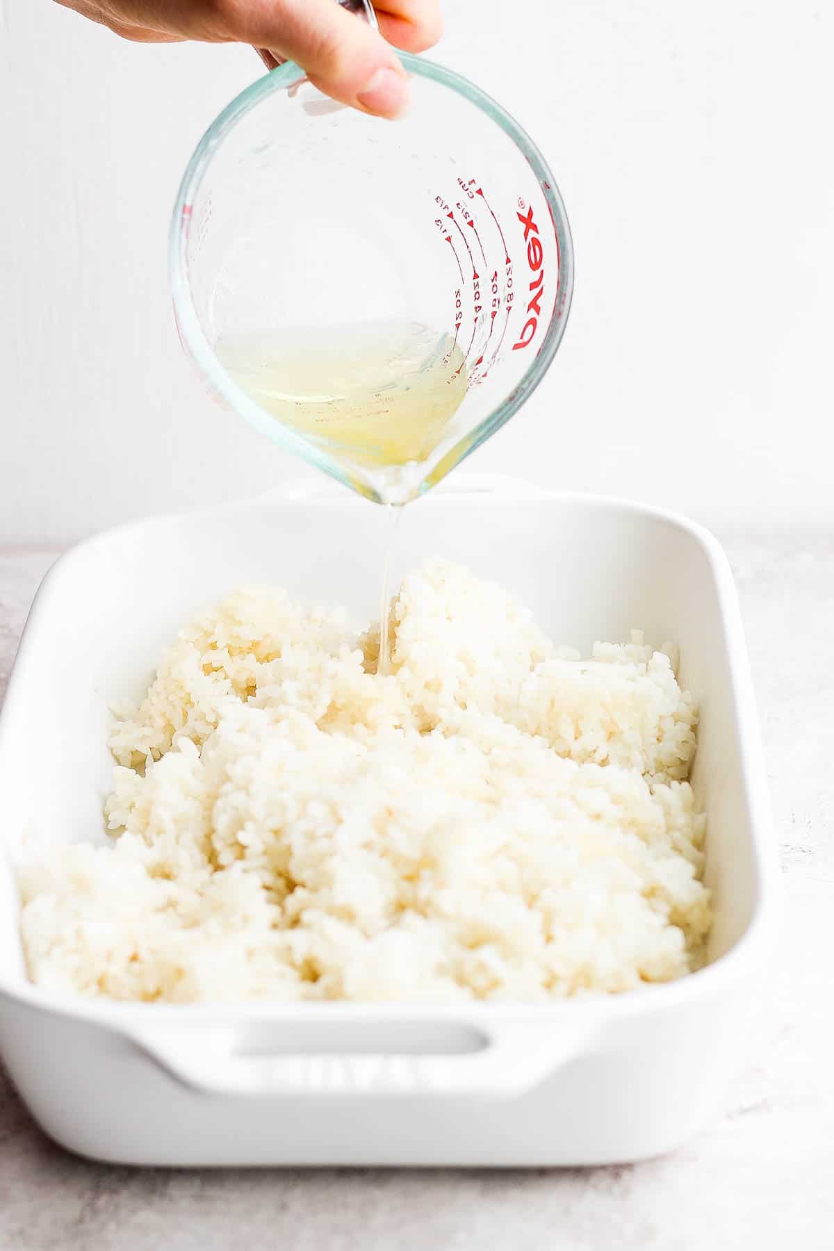 Rice vinegar mixture being poured over the cooked sushi rice in a white baking dish.