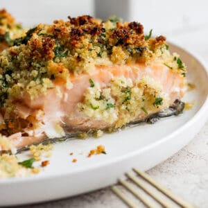 Side shot of a piece of panko crusted salmon on a white plate with the tip of a gold fork in the bottom right hand corner.