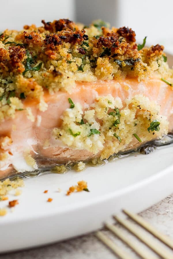 Side shot of a piece of panko crusted salmon on a white plate with the tip of a gold fork in the bottom right hand corner.