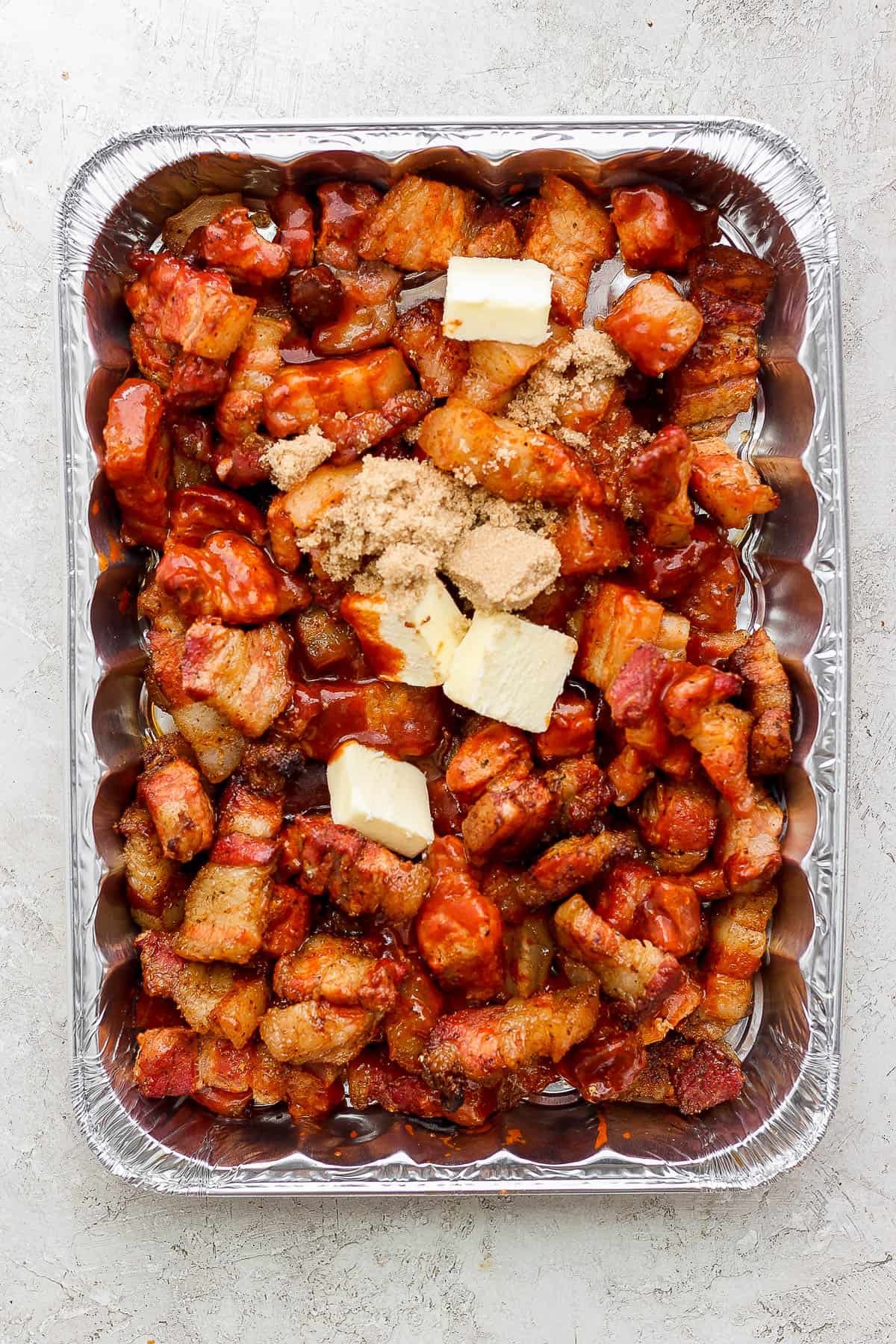 Pork belly pieces in an aluminum foil tray with butter, brown sugar, bbq sauce, and apple cider vinegar.