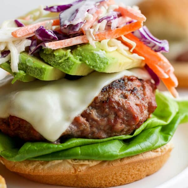 Close up of a pork burger with melted american cheese on a bun with lettuce, avocado and topped with a carrot and cabbage slaw and no top bun.