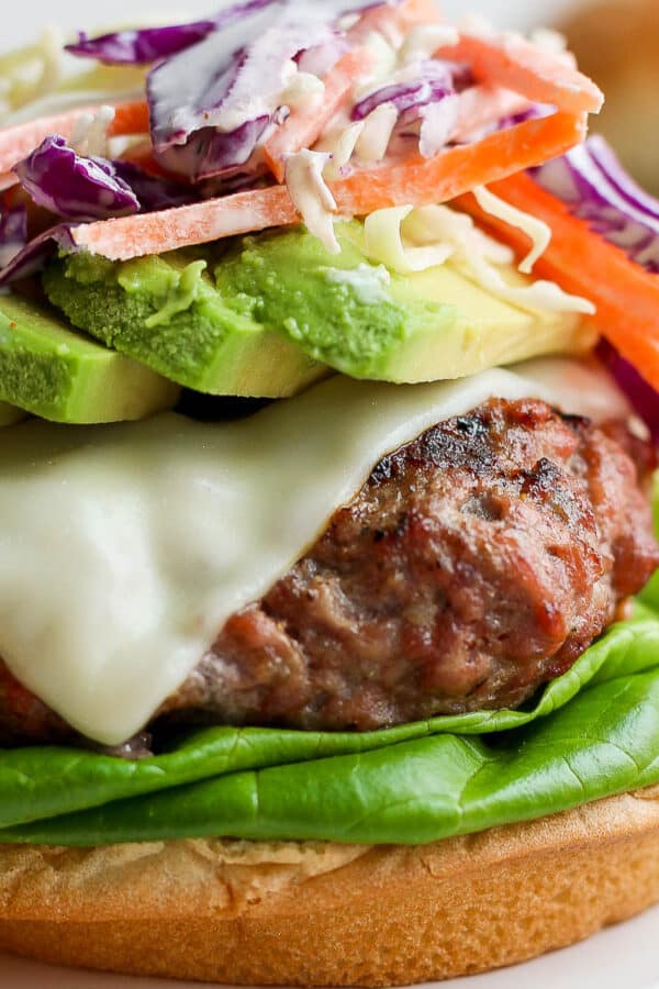 Close up of a pork burger with melted american cheese on a bun with lettuce, avocado and topped with a carrot and cabbage slaw and no top bun.
