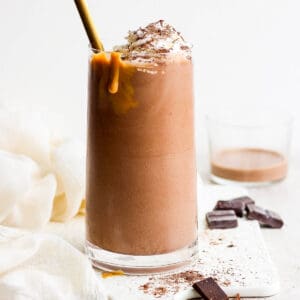Straight on shot of a chocolate protein shake in a glass with a drip of peanut butter sliding down the glass, whipped cream on top and a spoon sticking out.