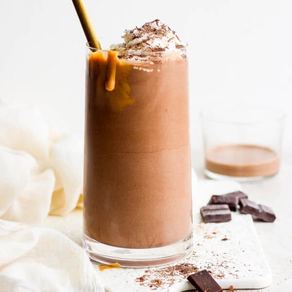 Straight on shot of a chocolate protein shake in a glass with a drip of peanut butter sliding down the glass, whipped cream on top and a spoon sticking out.