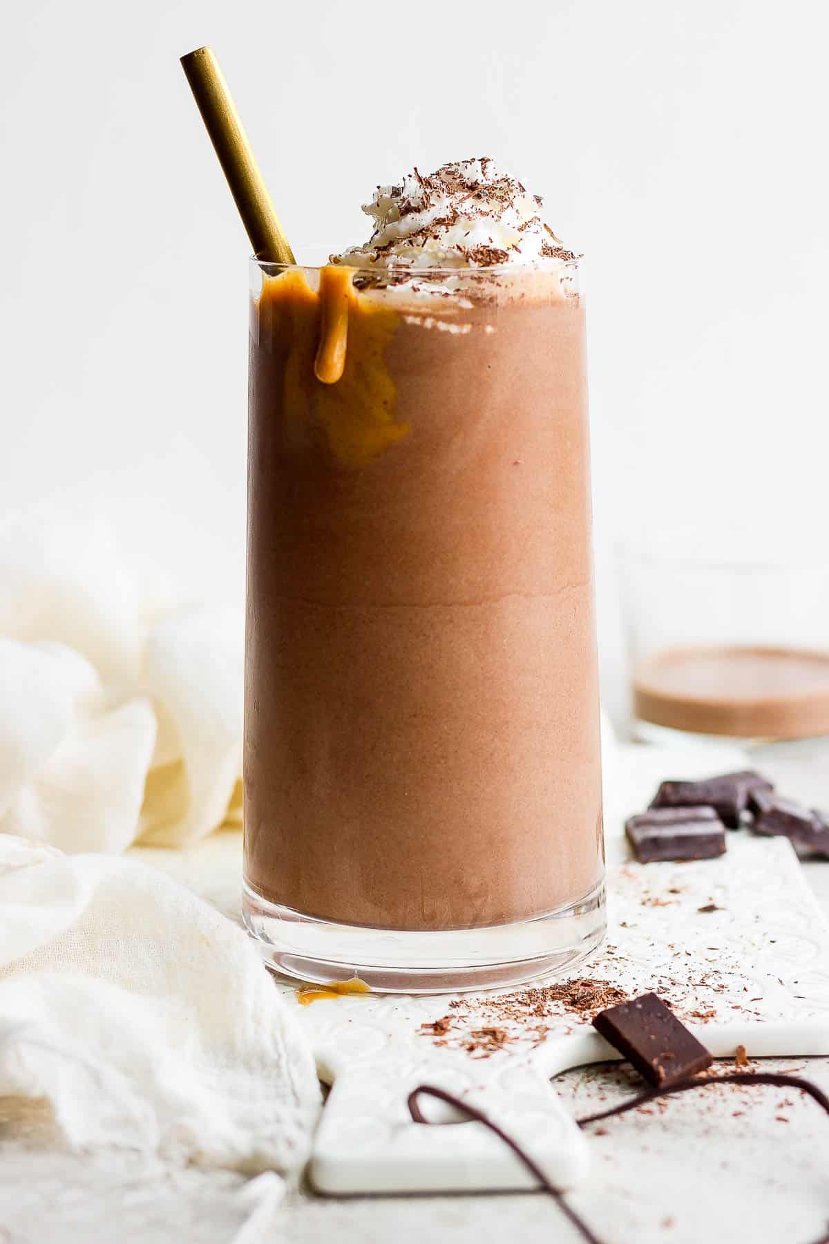 Chocolate protein shake in a glass topped with whipped cream and shaved chocolate with a golden straw.