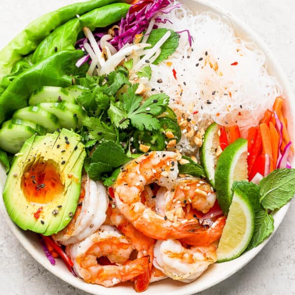 Top down shot of a shrimp spring roll bowl with avocado, cucumber, rice noodles, lettuce, limes and spring roll sauce on top.