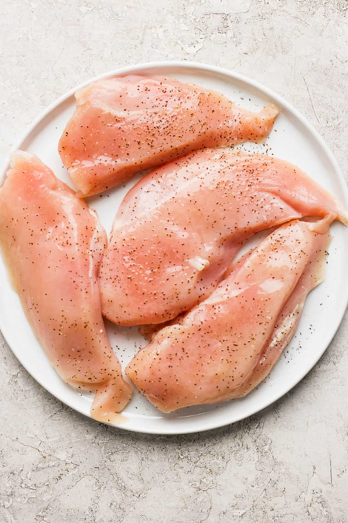 Raw chicken breasts on a white plate seasoned with salt & pepper.