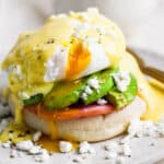 Straight on shot of an eggs benedict recipe with avocado and goat cheese crumbles and runny poached egg on a plate.