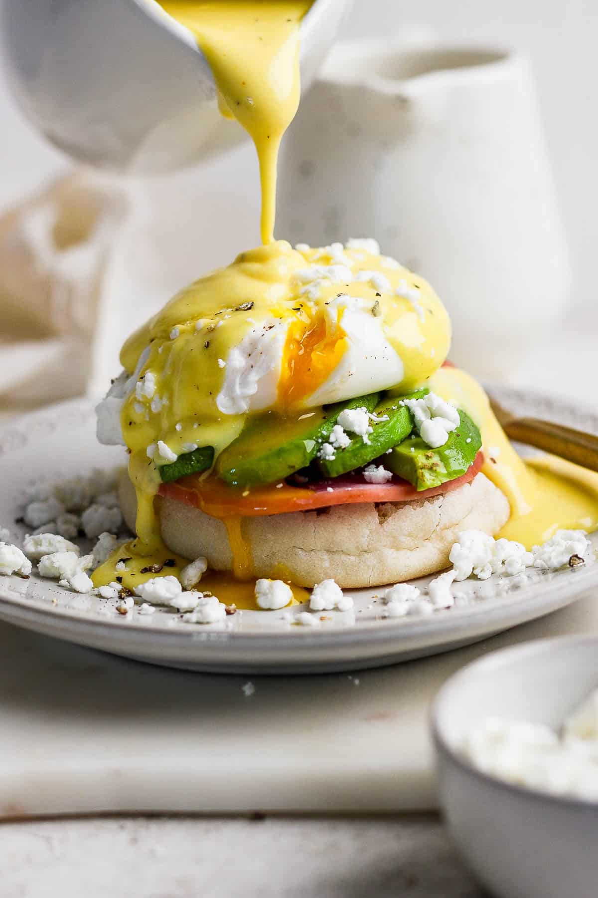 An assembled eggs Benedict with hollandaise sauce being poured on top.