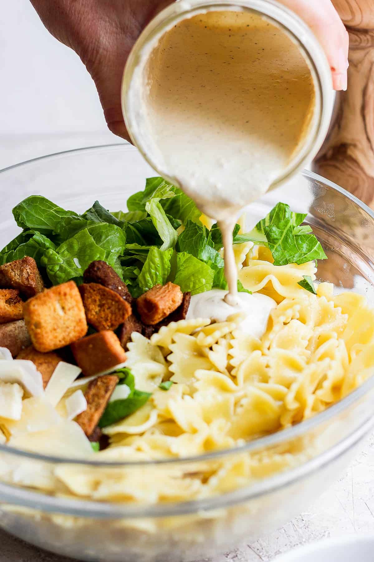 Caesar pasta salad ingredients in a bowl with dressing being poured on top.