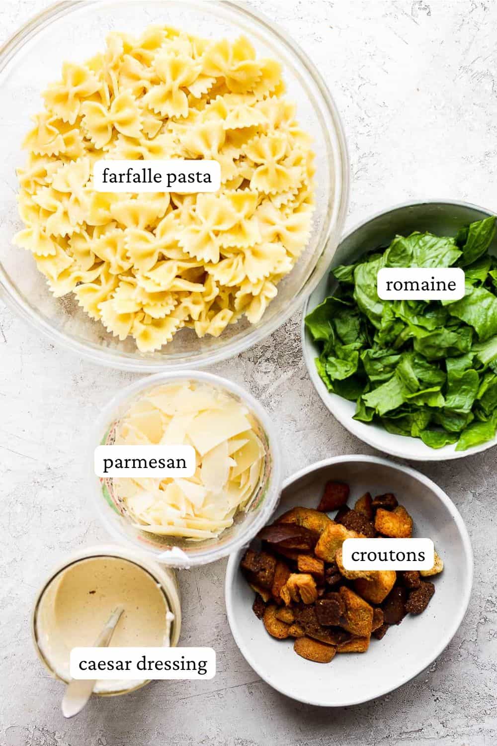 Ceasar pasta salad ingredients in individual bowls including farfalle pasta, romaine lettuce, parmesan cheese, croutons, and caesar dressing.