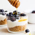 Straight on shot of a greek yogurt parfait in a short glass cup with crushed graham crackers on the bottom and then blueberries on top with honey being drizzled on top.