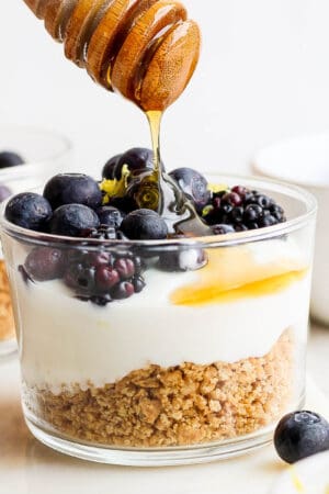 Straight on shot of a greek yogurt parfait in a short glass cup with crushed graham crackers on the bottom and then blueberries on top with honey being drizzled on top.