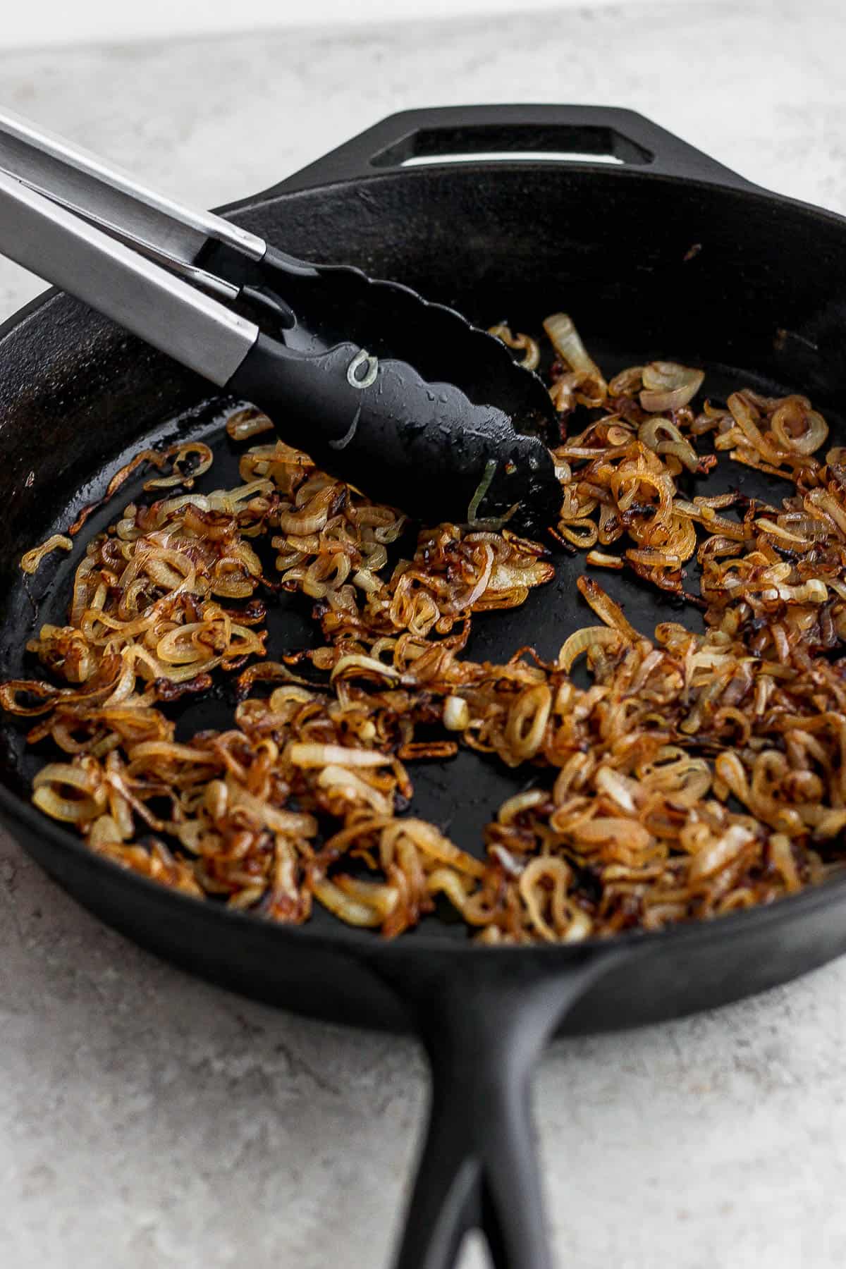 A pair of tongs moving crispy shallots around in a skillet.
