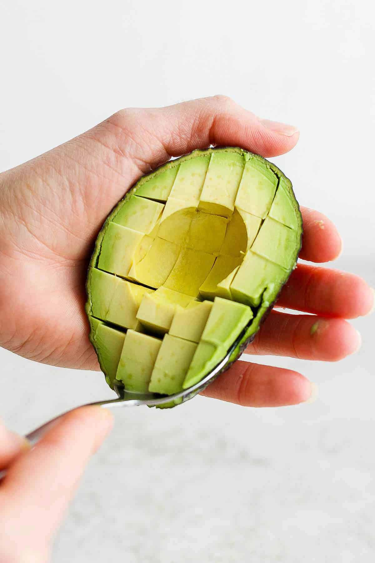 Two hands using a spoon to scoop avocado chunks out of the skin.