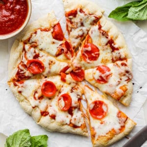 Top down shot of a grilled pizza cut into slices with pepperoni on top surrounded by a bowl of pizza sauce and fresh basil.