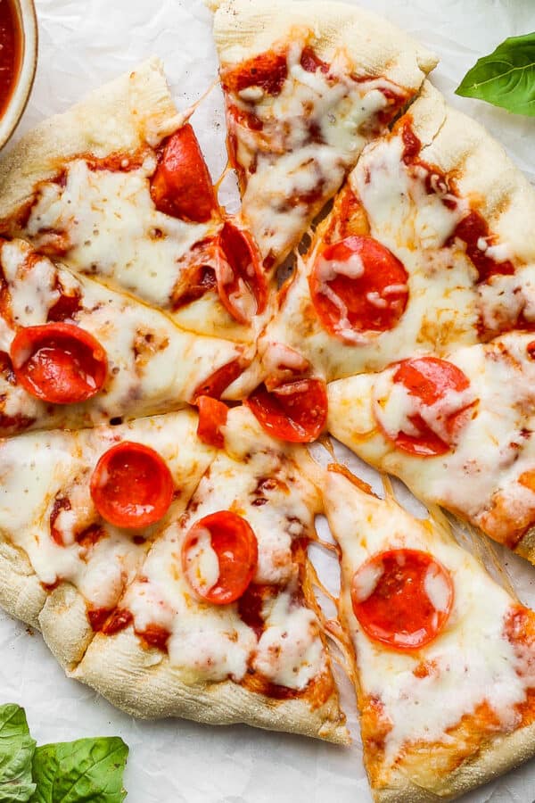 Top down shot of a grilled pizza cut into slices with pepperoni on top surrounded by a bowl of pizza sauce and fresh basil.